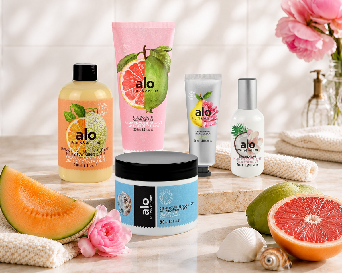 A promotional banner for Alo Essentials featuring several skincare and bath products including a yellow shampoo, a pink body wash, a blue scrub jar, and a white tube, styled with melon, grapefruit, and pink roses on a marble surface with French promotional text.